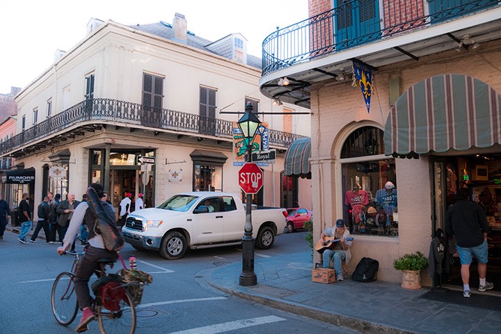 French Quarter intersection with bicyclist and driver negotiating right-of-way, serenaded by street musician sitting against the corner building