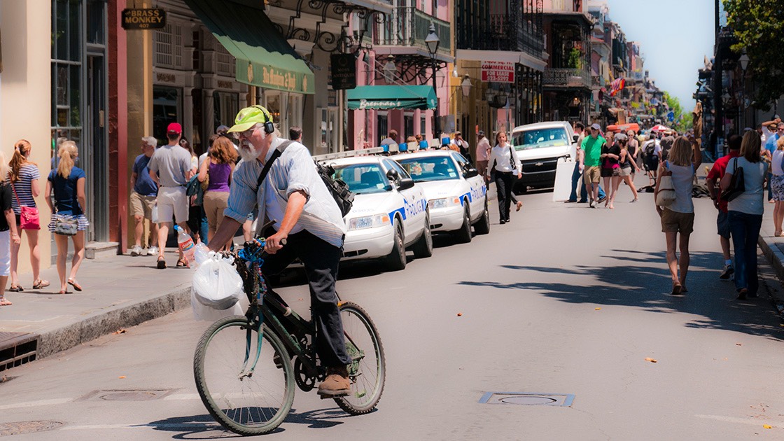 bicycle rider leaning into his turn off French Quarter street thronged by visitors, with green-sheathed cast iron galleries in the distance